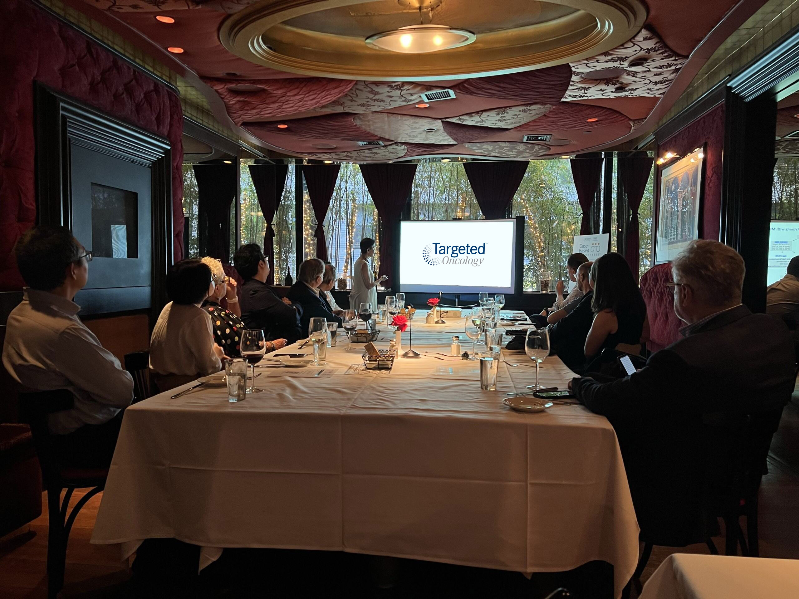 An elaborately decorated room with a long table. People are seated and facing a screen, possibly watching a presentation. The room has a luxurious feel.