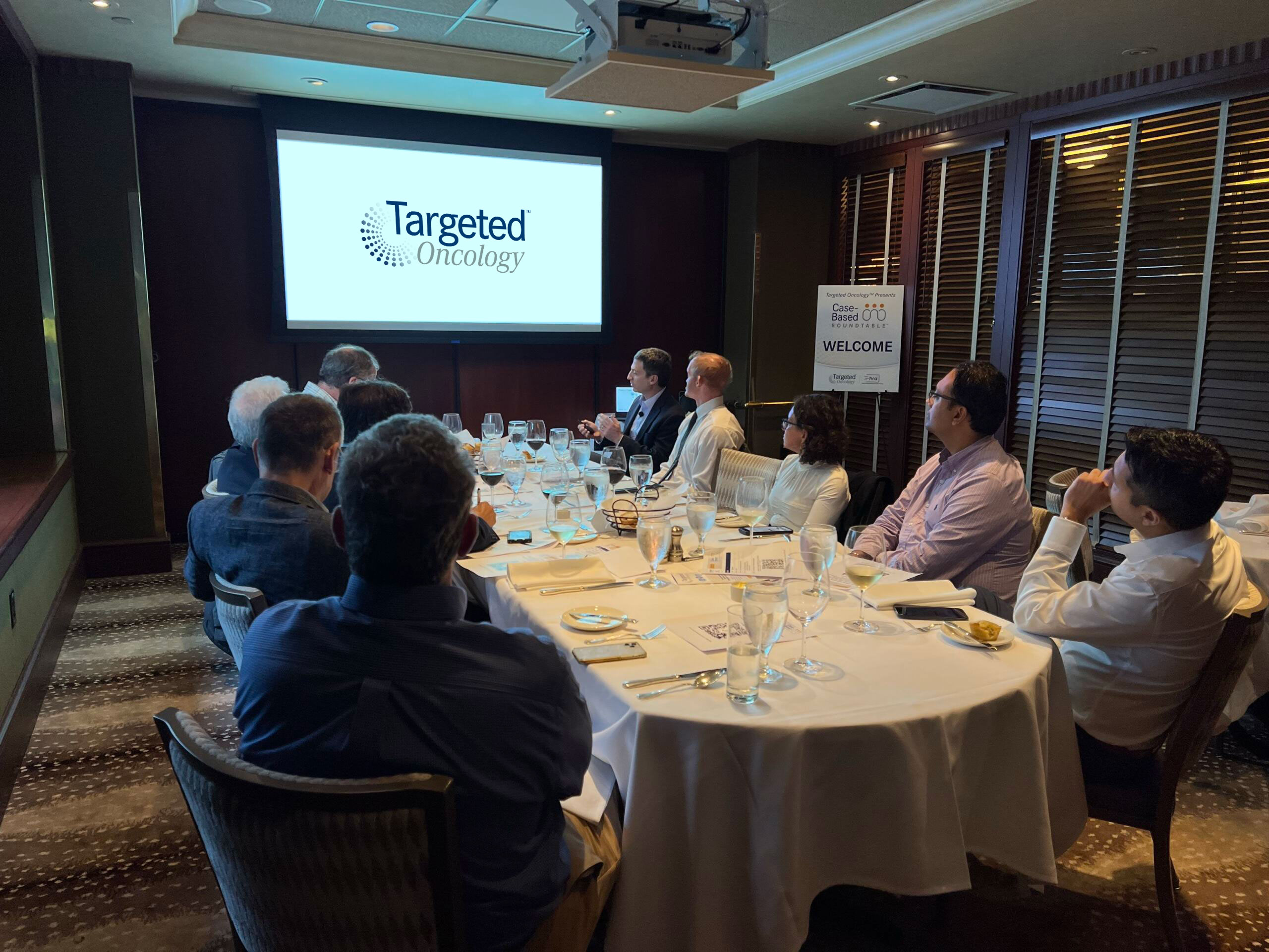 A Case-Based Roundtable event on Therapeutic Targets in CLL, guests are sitting around an oval table watching a presentation and engaging in discussion.