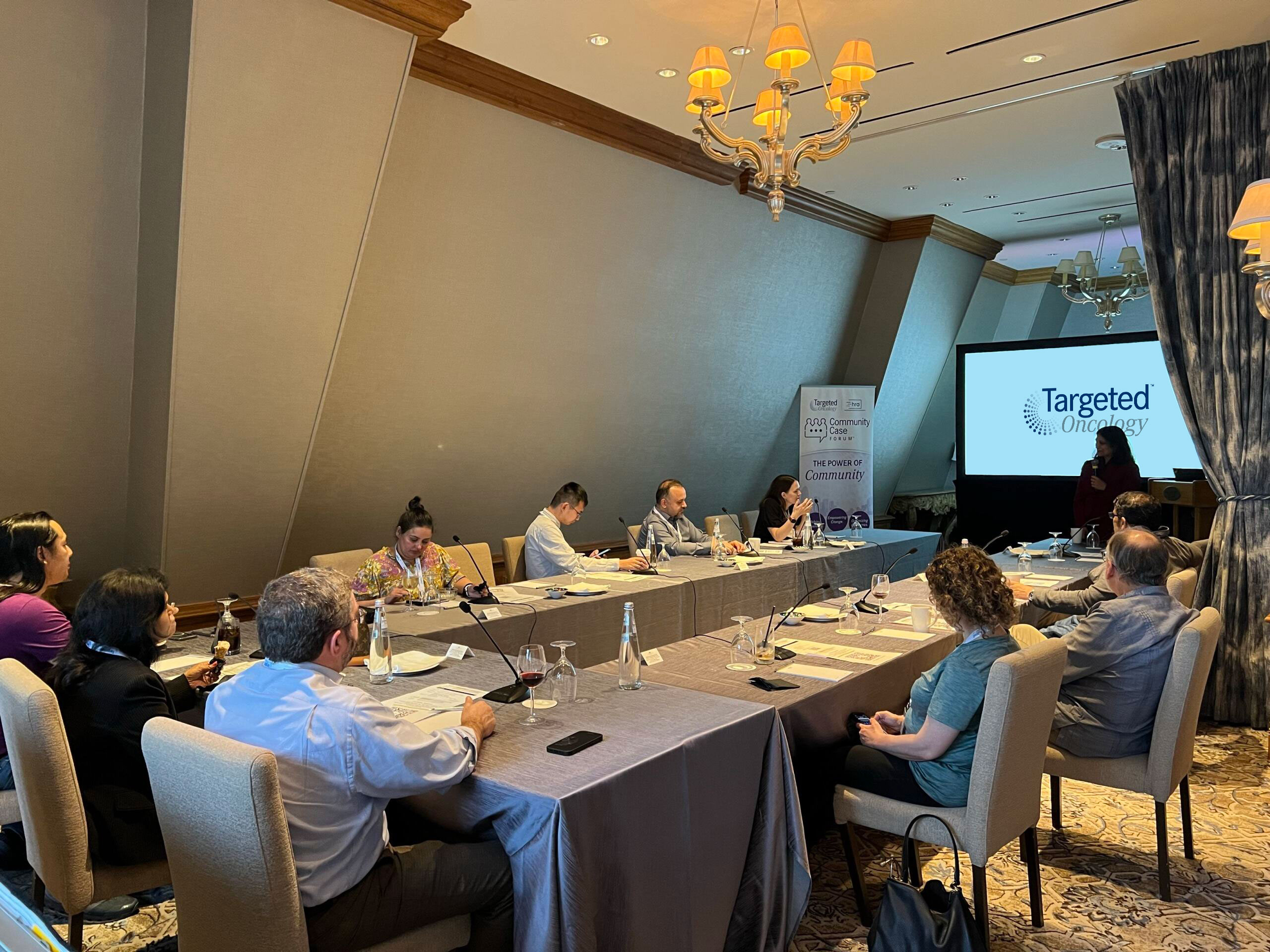 A large, elegant private dining room with several colored-clothed tables arranged in a U-shape. People are seated around the table, looking towards a large projection screen displaying a presentation.