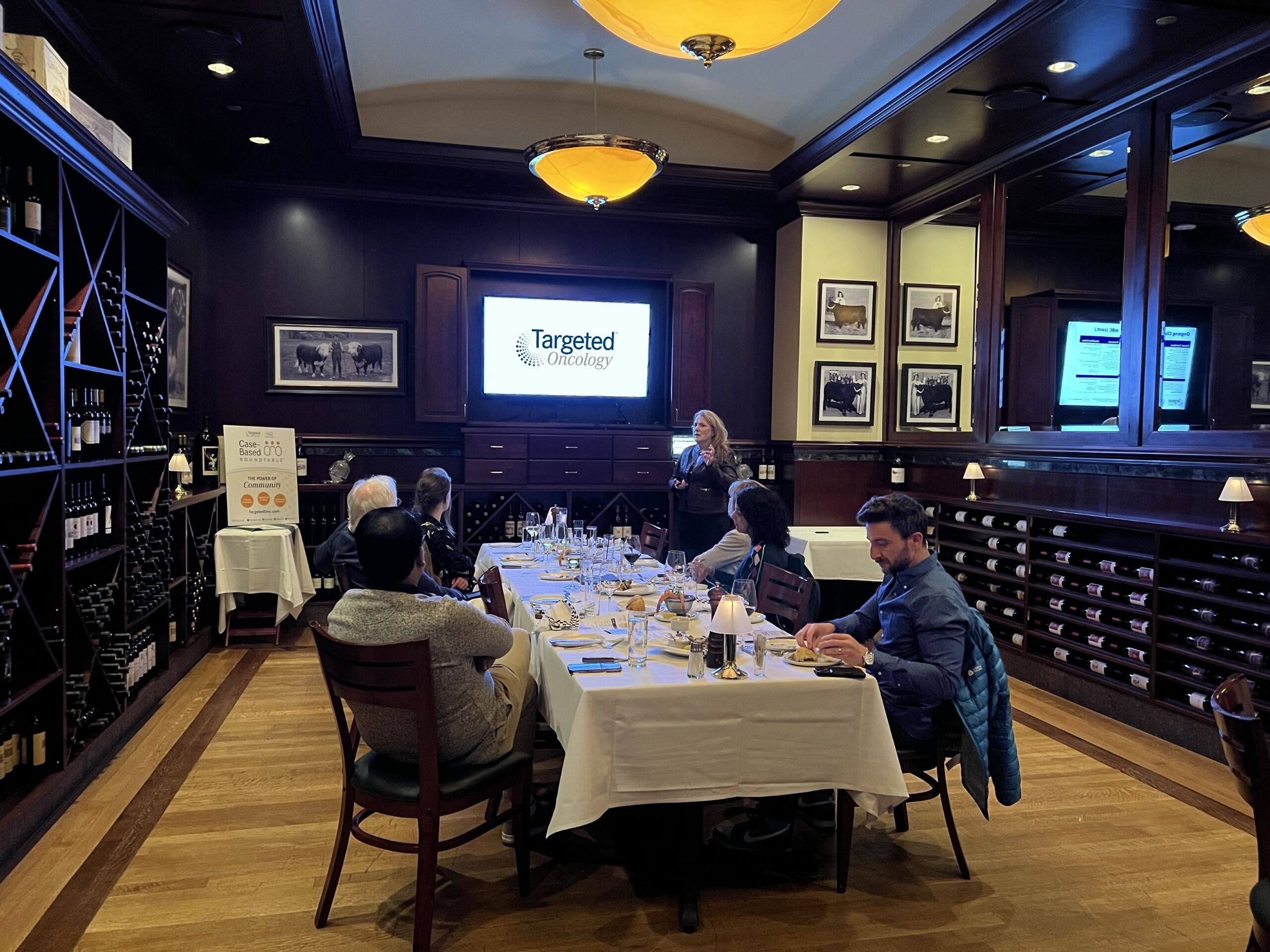 A Case-Based Roundtable event taking place in a small beautifully decorated room, a fancy wine shelf on the back wall, and guests are gathered at the table with drinks and watching the presentation on a screen in the front of the room beside a presentor standing at the podium.