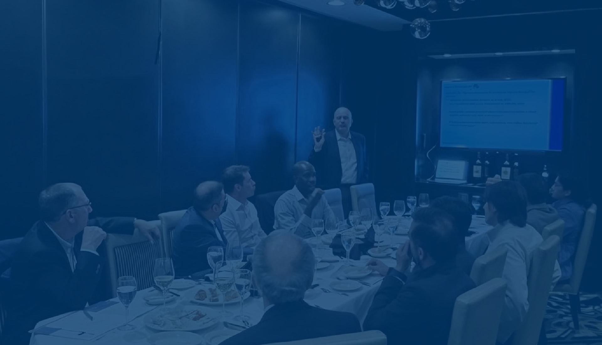 A group of people seated around a long dining table in a private event room, looking towards a large projection screen displaying a presentation. The tables are set with food and drinks. There is a presentor at the front of the room leading the presentation.